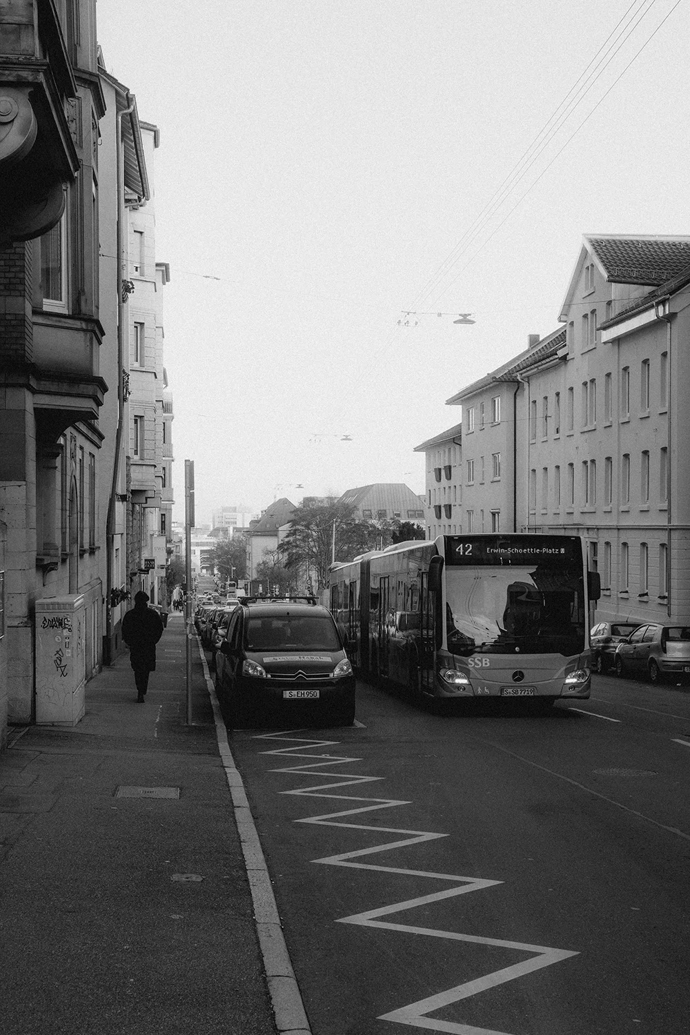 Eine belebte europäische Straßenansicht in Stuttgart. Historische Gebäude säumen die Straße, wo ein Bus parkt. Ein Zebrastreifen markiert den Vordergrund.