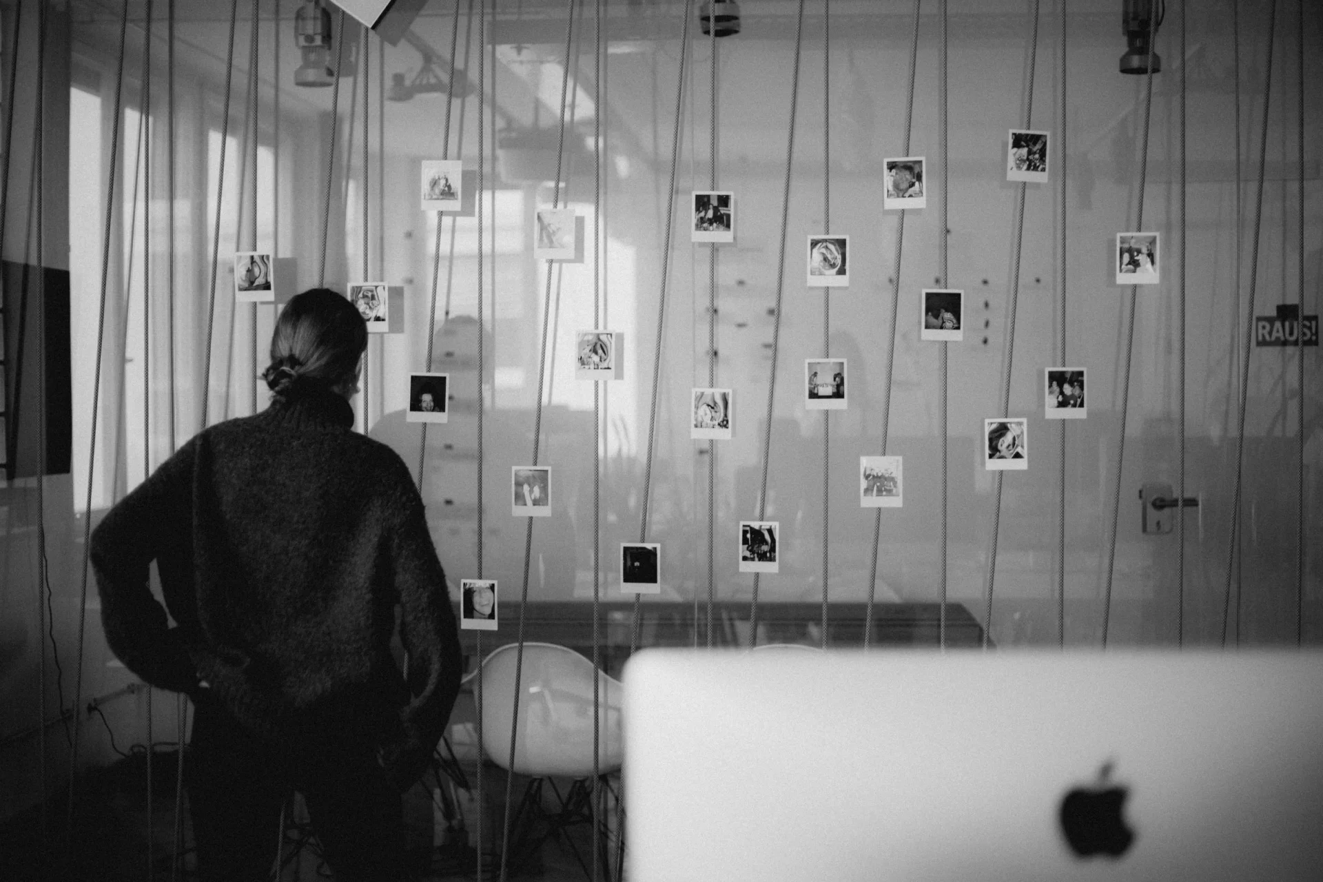 Monochrome Aufnahme eines hellen Innenraums. Eine Person steht vor großen Fenstern, neben einem Schreibtisch. Die Szene wirkt ruhig und akademisch.