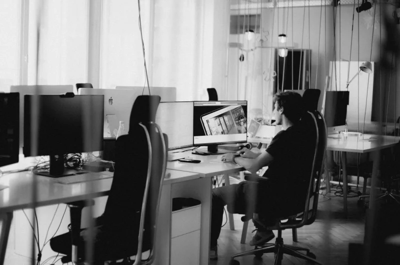 Ein Mann arbeitet konzentriert an einem Schreibtisch mit Monitoren und Büchern in einem hellen, professionellen Büro.